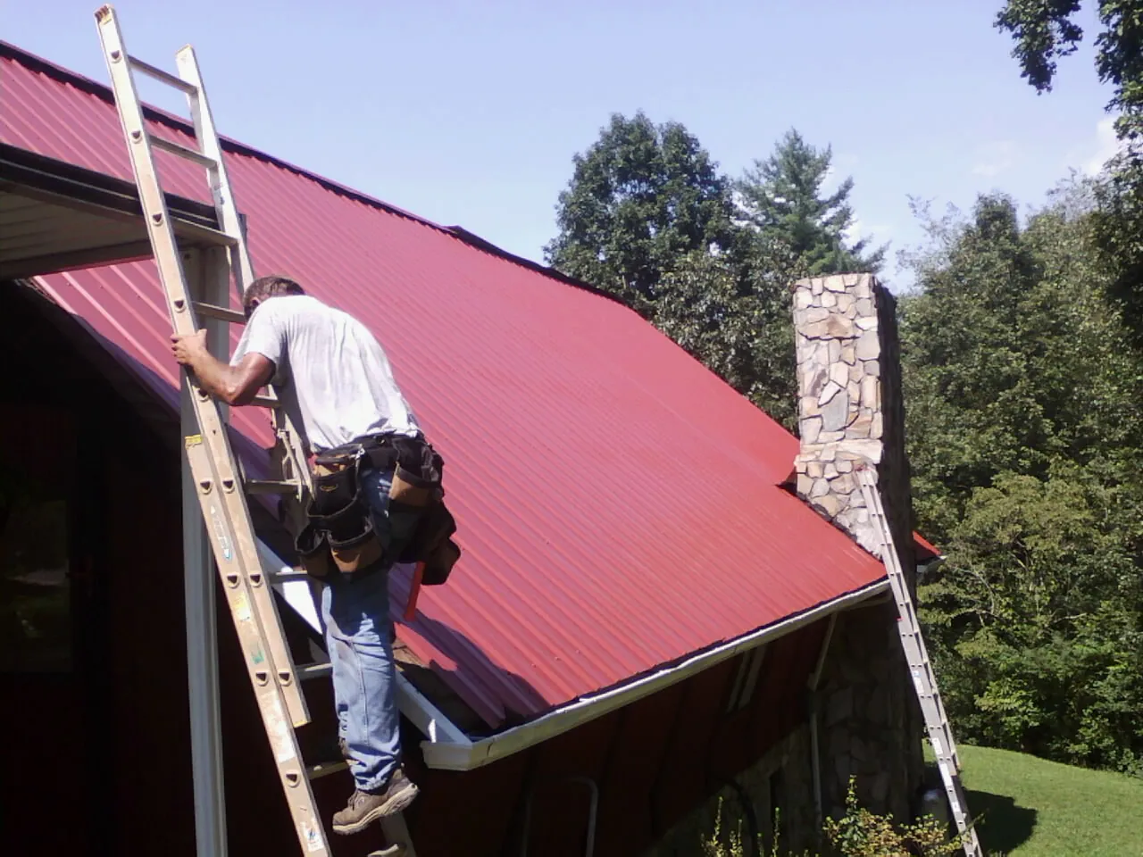 Roofer on a ladder beside a red metal roof and stone chimney — Soffit & Fascia Repair by Roofing Brothers in Morris Chapel